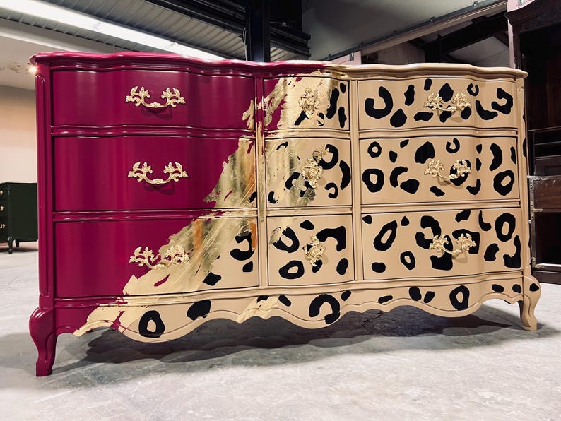 May include: A painted dresser with a unique design. The left side is a deep magenta, while the right side features a tan background with black leopard spots. Gold leaf accents and ornate hardware complete the look. The dresser has six drawers.