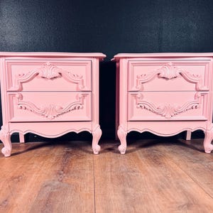 May include: Two matching pink nightstands with ornate carvings and two drawers each. The nightstands have curved legs and decorative molding. The background is a dark teal color, and the floor is a light brown wood.