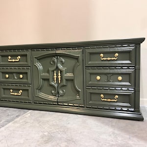 May include: A dark green dresser with gold-colored hardware. The dresser has multiple drawers and doors with ornate detailing. The hardware includes knobs and pulls. The dresser is a long, rectangular shape.