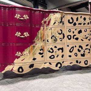 May include: A large, ornate dresser with a two-tone design. The left side is a deep magenta, while the right side is beige with black leopard spots. Gold leaf accents and ornate gold drawer pulls add a touch of luxury.