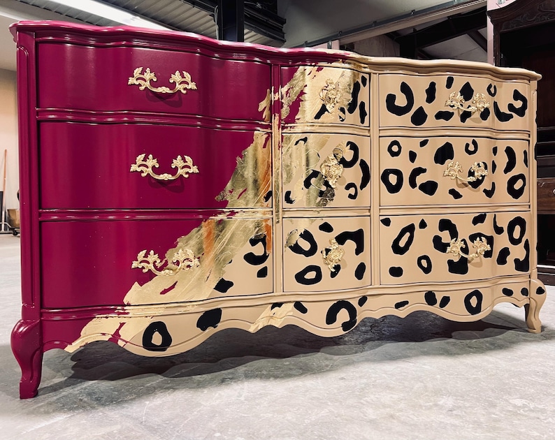 May include: A large, ornate dresser with a two-tone design. The left side is a deep magenta, while the right side is beige with black leopard spots. Gold leaf accents and ornate gold drawer pulls add a touch of luxury.