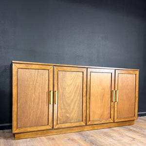 May include: A long wooden sideboard featuring four cabinet doors with a natural wood grain finish. Each door is framed with a raised border and equipped with a vertical, rectangular brass-colored metal handle. The unit has a flat top surface.