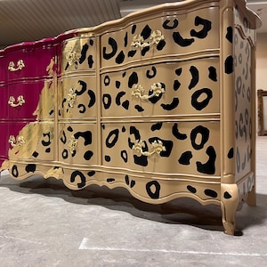 May include: A large, ornate dresser with a unique design. The left side is a deep magenta, transitioning to gold and tan with black leopard spots. Gold hardware and curved legs complete the look. A statement piece for any bedroom.