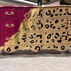 May include: A six-drawer dresser with a unique design. The left side is a deep magenta with gold hardware, while the right side features a tan leopard print. Gold leaf accents the transition between the two sides. The dresser has curved legs.