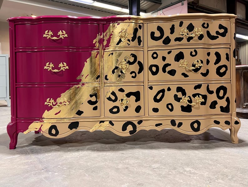 May include: A six-drawer dresser with a unique design. The left side is a deep magenta with gold hardware, while the right side features a tan leopard print. Gold leaf accents the transition between the two sides. The dresser has curved legs.