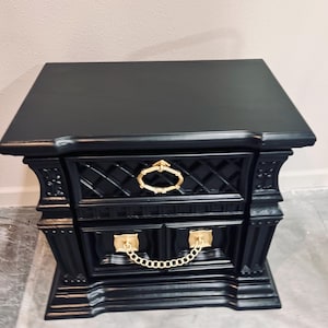 May include: A black nightstand with gold-colored hardware. The nightstand has two drawers and a top surface. The drawer pulls are gold-colored rings and the drawer fronts have a diamond pattern. The nightstand is approximately 24 inches wide, 18 inches deep, and 28 inches tall.