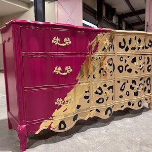 May include: A painted dresser with three drawers on each side. The left side is magenta, the right side is tan with black leopard spots. Gold leaf and gold drawer pulls add detail.