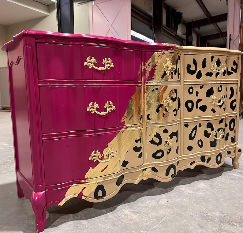 May include: A painted dresser with three drawers on each side. The left side is magenta, the right side is tan with black leopard spots. Gold leaf and gold drawer pulls add detail.