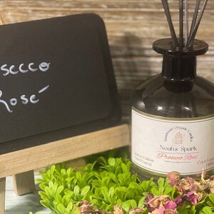 May include: A glass bottle diffuser with a black lid and a white label that reads "Noah's Spark Prosecco Rose" with a small illustration of a church. The bottle is surrounded by dried rose petals and green foliage.