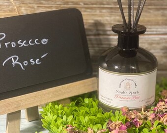 May include: A glass bottle diffuser with a black lid and a white label that reads "Noah's Spark Prosecco Rose" with a small illustration of a church. The bottle is surrounded by dried rose petals and green foliage.