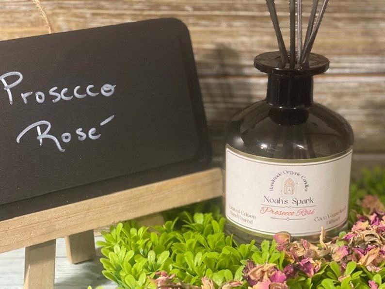 May include: A glass bottle diffuser with a black lid and a white label that reads "Noah's Spark Prosecco Rose" with a small illustration of a church. The bottle is surrounded by dried rose petals and green foliage.