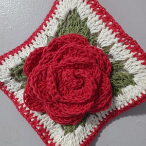May include: A crocheted square with a red rose in the center. The square is bordered with white and green crocheted trim.