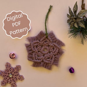 Peut inclure: Un modèle numérique PDF pour une décoration de Noël en macramé en forme de flocon de neige violet. Le flocon de neige est suspendu à une ficelle verte et a une frange autour des bords.