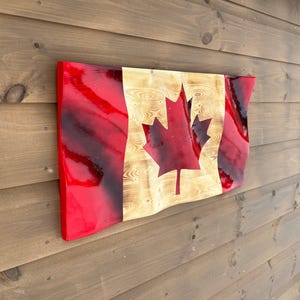 May include: A wooden Canadian flag with a red border and a maple leaf emblem. The flag has a wavy design and is mounted on a wooden wall. The maple leaf is a darker shade of red.