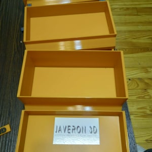 Może przedstawiać: Pomarańczowe prostokątne pudełka do przechowywania, ułożone w stos i otwarte, z błyszczącym wykończeniem. Pudełka są puste, odsłaniając wnętrze. Na dolnym pudełku znajduje się biała etykieta z napisem "JAVERON 3D CUSTOM PRINTING AND DESIGN".