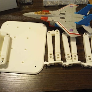 Op de afbeelding: Een witte plastic basis met meerdere gaten en zes witte, scharnierende plastic armen. Een grijze en blauwe speelgoed straaljager staat op de basis.