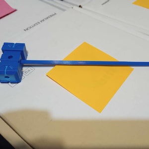 Puede incluir: Un martillo de plástico azul con un mango azul. La cabeza del martillo está hecha de tres cubos conectados. El mango está unido al cubo central. El martillo está sobre una superficie blanca con una nota adhesiva amarilla.