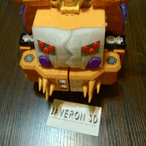 May include: A close-up of a yellow and white toy robot with orange horns and a white face. The robot has red and purple accents. A business card with the text "JAVERON 3D CUSTOM PRINTING AND DESIGN" is in the foreground.