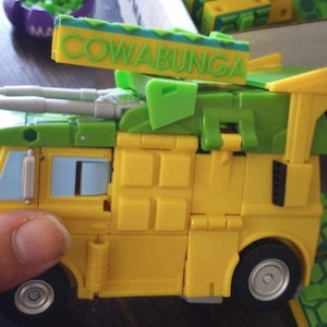 May include: A yellow toy truck with a green roof and a green sign that says "Cowabunga!" The truck has a silver cannon on the front and a silver wheel on the side.