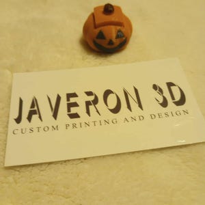May include: A small, orange 3D-printed pumpkin with a carved face and a stem. The pumpkin is sitting on a soft, cream-colored surface. A white card with the text "JAVERON 3D CUSTOM PRINTING AND DESIGN" is in the foreground.