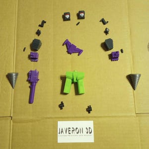 May include: A collection of 3D-printed toy components in shades of purple, green, and black, displayed on a cardboard background. The parts include robot heads, legs, and other elements. Two grey drill bits are also present, along with a white label reading "JAVERON 3D CUSTOM PRINTING AND DESIGN."