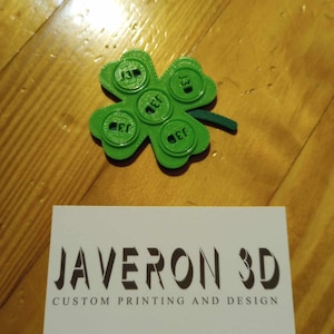 Op de afbeelding: Een groene klaver met vier bladeren met vier ronde knoppen waarop op elk "1e" staat. De klaver ligt op een houten oppervlak met een visitekaartje waarop "JAVERON 3D AANPASSINGSDRUK EN -ONTWERP" staat.