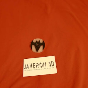 Puede incluir: Un pin redondo con el logotipo de Batman en negro y amarillo. El pin está sobre una superficie roja. Debajo, una tarjeta de visita blanca con el texto "JAVERON 3D CUSTOM PRINTING AND DESIGN".