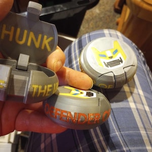 Może przedstawiać: Trzy szare, okrągłe pojemniki wydrukowane w 3D z różnymi wzorami. Pierwszy pojemnik ma napis "HUNK" wydrukowany na żółto. Drugi pojemnik ma wzór wilka w kolorze żółtym i niebieskim. Trzeci pojemnik ma napis "DEFENDER OF" wydrukowany na czerwono.