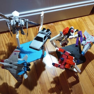 Peut inclure: Une collection de cinq figurines articulées et un modèle de véhicule présentés sur des socles. L'ensemble comprend un robot oiseau gris, une voiture de sport argentée, un robot blanc et rouge, un avion de chasse gris et bleu, et un robot rouge et bleu.