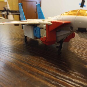 Puede incluir: Un avión de juguete blanco, rojo y azul con una cabina amarilla. El avión tiene un tren de aterrizaje retráctil y está en posición de aterrizaje.