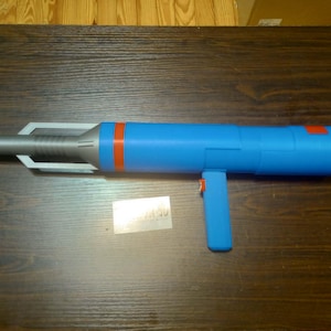 May include: A blue and silver toy blaster with a long cylindrical barrel, a vertical handle, and a bright orange ring accent near the front. The blaster features a metallic silver muzzle section with a geometric frame and a solid blue main body.