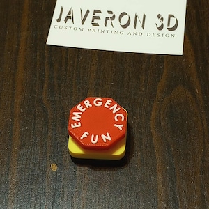 Może przedstawiać: Przedmiot drukowany w 3D, z czerwonym ośmiokątem z białym napisem "EMERGENCY FUN" na żółtej kwadratowej podstawie. Przedmiot znajduje się na ciemnej powierzchni z fakturą drewna, z białą etykietą nad nim.