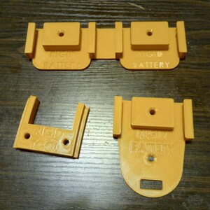 Könnte beinhalten: Drei orangefarbene, 3D-gedruckte Batteriehalter mit dem Text "Rigid Battery" auf jedem. Die Halter sind so konzipiert, dass sie Batterien an ihrem Platz halten.