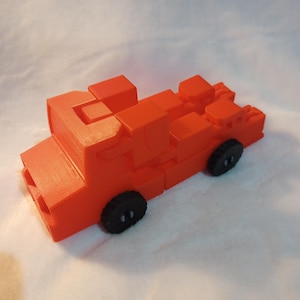 Pode incluir: Um caminhão de bombeiros de brinquedo vermelho feito de blocos de construção. O caminhão tem rodas pretas e é feito de plástico liso.