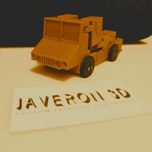 Pode incluir: Um pequeno caminhão de brinquedo de plástico cor de areia com uma caçamba basculante. O caminhão tem uma base e rodas de plástico preto. O caminhão é impresso com o texto "JAVERON 3D CUSTOM PRINTING AND DESIGN".