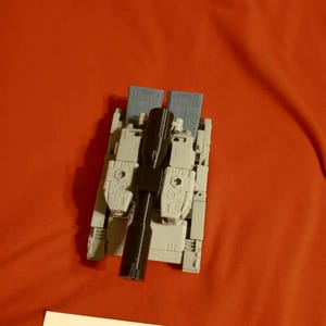 Pode incluir: Um tanque de brinquedo cinza e preto com um cano preto, sobre um fundo vermelho. O tanque tem painéis retangulares cinza e um componente central preto. O texto "JAVERON 3D" é visível em um rótulo branco.