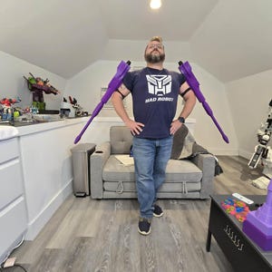 May include: A navy blue t-shirt featuring a white graphic of a robotic face and the text MAD ROBOT. The shirt is worn with two long, purple, pointed robotic arm attachments extending outward from the shoulders, secured by black elastic bands.