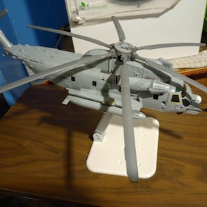 Puede incluir: Un helicóptero de juguete gris con un soporte blanco. El helicóptero tiene un cuerpo largo y delgado y cuatro aspas. El soporte es simple y tiene una base rectangular.