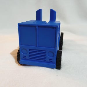 Pode incluir: Um caminhão de brinquedo azul com rodas pretas e uma cabine elevada. O caminhão tem uma grade e faróis.