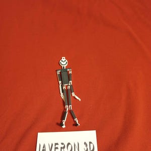 May include: A small, articulated figure with a white head and a smiling face, black and white striped body, and white limbs. The figure is standing on a red surface. A white card with the text "JAVERON 3D CUSTOM PRINTING AND DESIGN" is in the foreground.