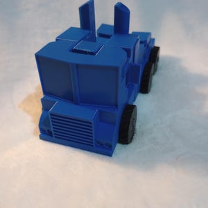 Pode incluir: Um caminhão de brinquedo azul com uma cabine elevada e dois tubos de escape verticais. O caminhão tem rodas pretas e uma grelha frontal detalhada.