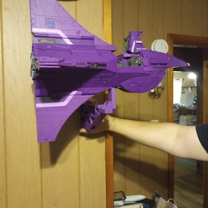 Op de afbeelding: Een paarse speelgoedjet met witte accenten, gemonteerd aan een muur. De jet heeft een grote staartvin en vleugels, met zichtbare motordetails. Het ontwerp suggereert een futuristisch of sciencefiction thema. Het speelgoed is waarschijnlijk van plastic.