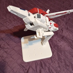Op de afbeelding: Een wit en rood modelvliegtuig met een standaard. Het model heeft een gedetailleerd ontwerp met vleugels en een cockpit. De standaard is wit en rechthoekig met verschillende gaten. Het vliegtuig is een speelgoed of verzamelobject.