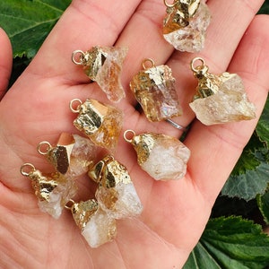 May include: A collection of eight gold-plated, raw, clear quartz crystal pendants. The crystals are various sizes and shapes, with some having a slight yellow tint.