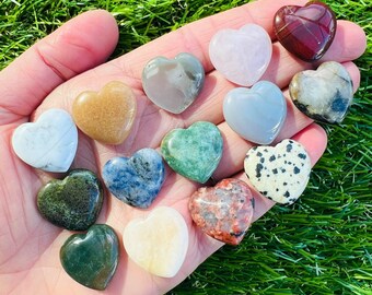 Può includere: Una collezione di 14 pietre levigate a forma di cuore in vari colori e motivi, tra cui verde, rosso, marrone, bianco e blu. Le pietre sono disposte su una mano, con le punte delle dita visibili.