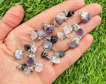 Può includere: Una collezione di piccoli ciondoli di pietre preziose grezze in vari colori, tra cui viola, nero e trasparente. Ogni pietra è sormontata da un cappuccio in metallo argentato e ha un piccolo anello per il fissaggio a una catena. I ciondoli sono esposti su una mano.