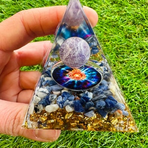May include: A clear crystal pyramid with a purple amethyst sphere at the top. The pyramid is filled with blue and white stones and gold flakes. The base of the pyramid is surrounded by green grass.