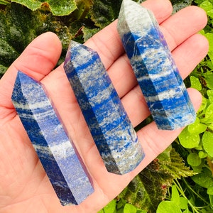 May include: Three blue and white lapis lazuli crystal points, also known as lapis lazuli wands, are displayed on a hand. The points are faceted and have a natural, raw look.