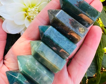 May include: Six green and brown moss agate points stacked on top of each other in a hand. The points are polished and have a smooth, glassy finish.