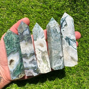 May include: Five moss agate crystal towers with varying patterns of green and white. The polished stones are held in a hand against a backdrop of green grass. Each tower has a unique shape and internal structure.
